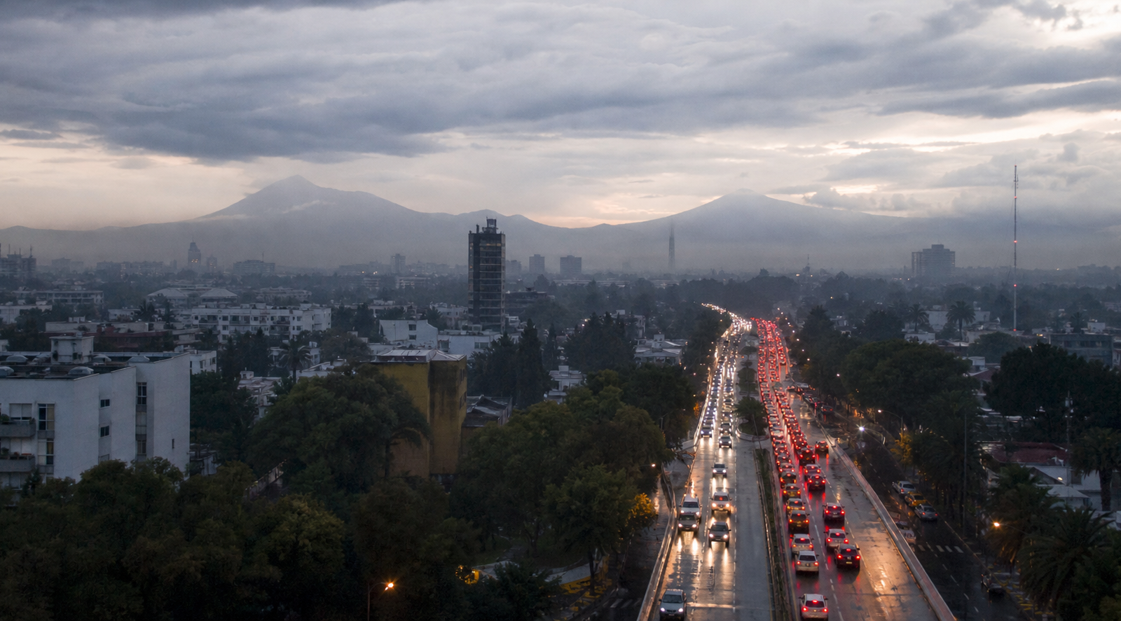 Tráfico vehicular en una avenida de la Ciudad de México al amanecer, con pavimento mojado y cielo nublado.