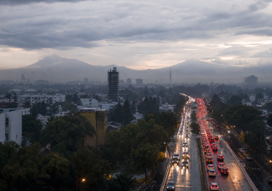 Tráfico vehicular en una avenida de la Ciudad de México al amanecer, con pavimento mojado y cielo nublado.