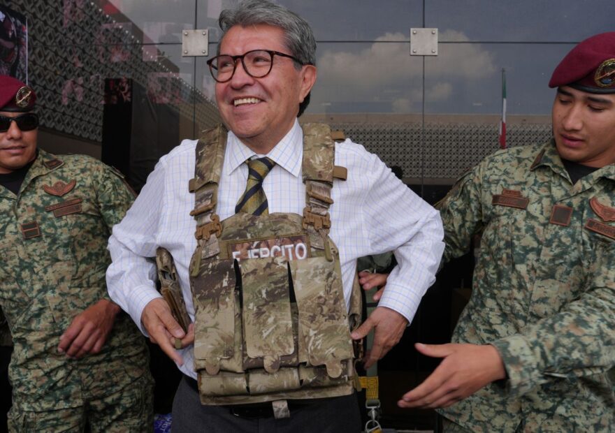 Ricardo Monreal prueba un chaleco táctico acompañado de dos militares durante una exhibición.