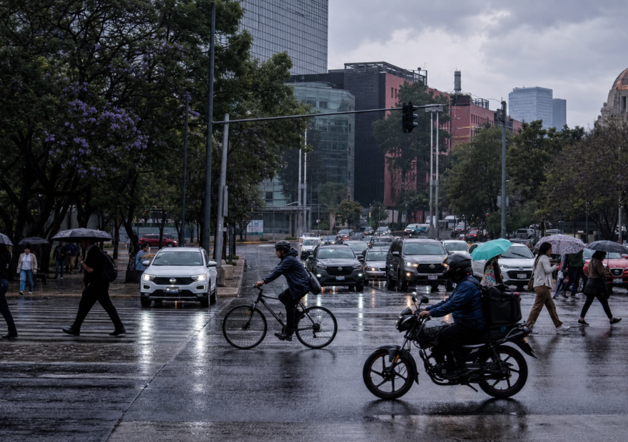 Automóviles avanzan lentamente sobre una avenida de la Ciudad de México mientras peatones cruzan con paraguas bajo un cielo nublado; al fondo se observan señales viales y edificios urbanos.
